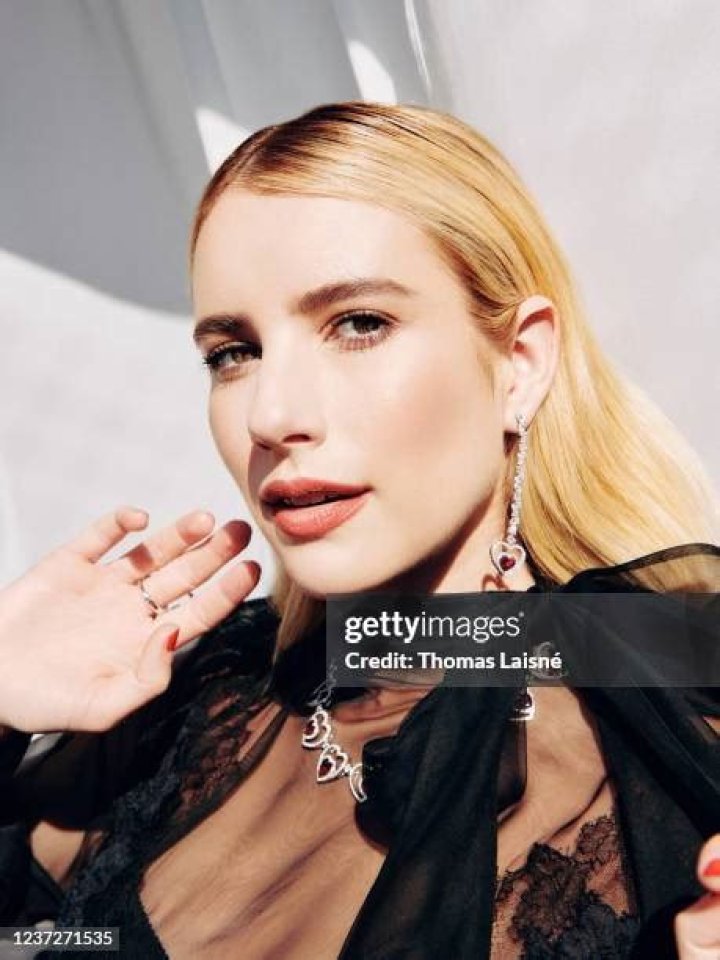 Emma Roberts poses holding a Barbie next to shelf of dolls and collage on mirror in her house