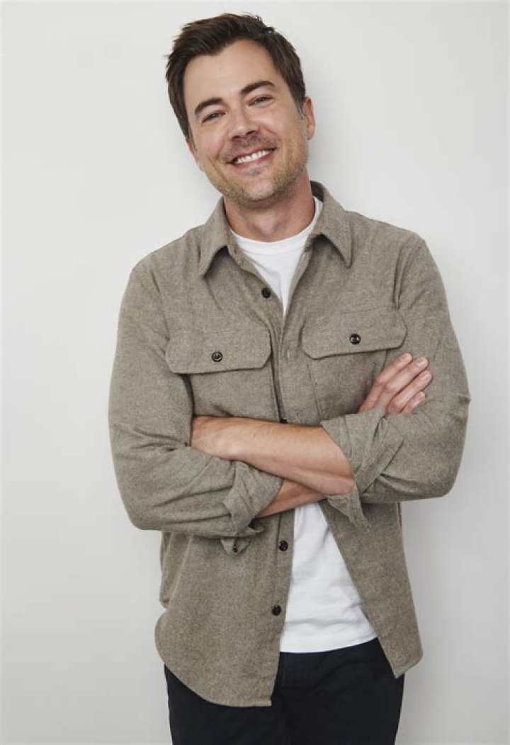 Matt Long in a red jacket and grey cap in front of a lake and hill.
